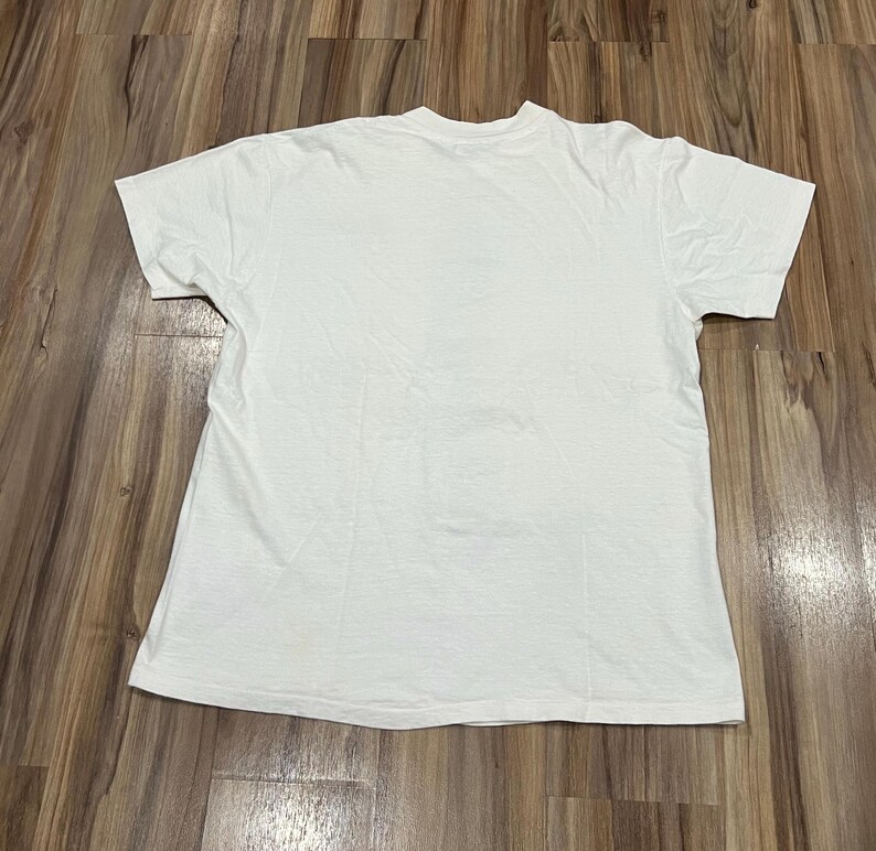 May include: A plain white t-shirt with short sleeves, laid flat on a wooden surface. The shirt is made of a soft, likely cotton material. The t-shirt is a basic, versatile garment.