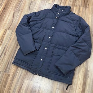 Puede incluir: Una chaqueta acolchada azul oscuro con cierre de botones y un bolsillo grande en la parte delantera. La chaqueta tiene un diseño acolchado y un cuello alto.