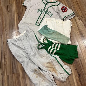 May include: A gray baseball jersey with green trim and the word Padres, a white t-shirt, a light green long-sleeve shirt, gray baseball pants with green side stripes and brown stains, and a green and white striped ribbed tank top.