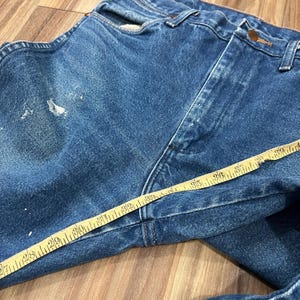 Pu&ograve; includere: Jeans in denim blu con lavaggio medio, chiusura a bottoni e schizzi di vernice bianca sul tessuto. Un metro a nastro giallo &egrave; posizionato sulla parte anteriore, mostrando una misura di circa 99 cm lungo la zona della vita.