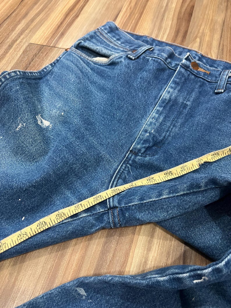 Pu&ograve; includere: Jeans in denim blu con lavaggio medio, chiusura a bottoni e schizzi di vernice bianca sul tessuto. Un metro a nastro giallo &egrave; posizionato sulla parte anteriore, mostrando una misura di circa 99 cm lungo la zona della vita.
