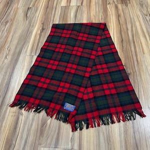 May include: A rectangular scarf featuring a red, dark green, and navy blue plaid pattern. The scarf has a fringed edge on both ends and includes a small blue rectangular label with white text that reads Pendleton, Warranted to be a Pendleton.