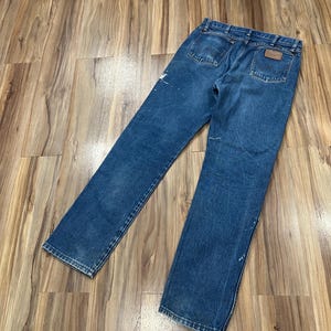 Pu&ograve; includere: Un paio di jeans in denim blu con un classico design a cinque tasche, una toppa in pelle marrone sulla tasca posteriore destra, passanti per cintura e dettagli con schizzi di vernice bianca sul tessuto, visti da una prospettiva posteriore.