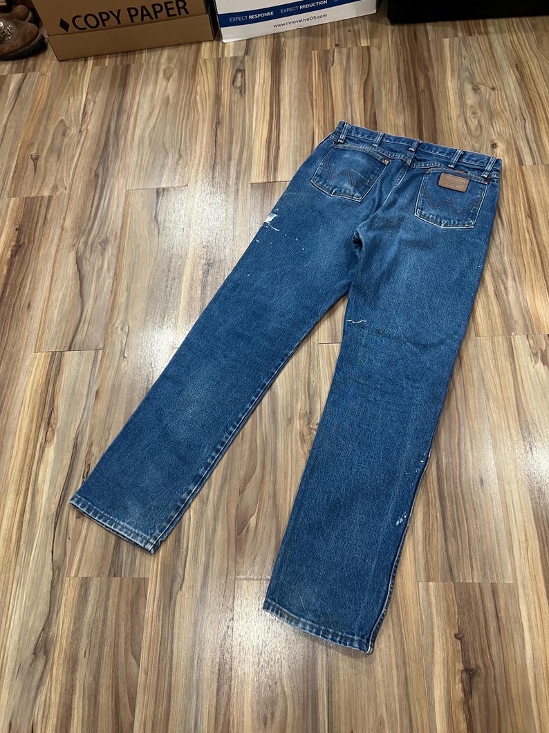 Pu&ograve; includere: Un paio di jeans in denim blu con un classico design a cinque tasche, una toppa in pelle marrone sulla tasca posteriore destra, passanti per cintura e dettagli con schizzi di vernice bianca sul tessuto, visti da una prospettiva posteriore.