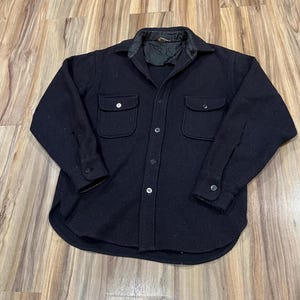 May include: A solid black long-sleeve shirt jacket featuring a button-down front, a pointed collar, and two chest pockets with buttoned flaps. The garment is constructed from a thick, textured fabric and includes a black quilted inner lining.