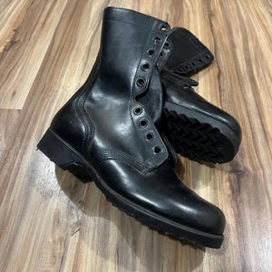 May include: Pair of black leather combat boots with multiple eyelets for laces. The boots have a high-top design and a thick, textured sole. The boots are displayed on a wooden surface.