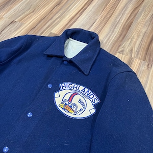 Pode incluir: Uma jaqueta de beisebol azul marinho com um remendo de feltro no peito esquerdo. O remendo é branco com detalhes azuis e vermelhos e diz "HIGHLANDS BIRDS".