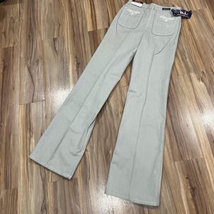 May include: Light gray flared pants with white embroidered details on the back pockets. The pants are made of a soft material and have a high waist. The pants are laid out on a wooden surface.