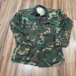 Könnte beinhalten: Tarnmusterjacke mit mehreren Taschen und Knopfverschlüssen. Die Jacke hat ein grünes, braunes und schwarzes Waldtarnmuster. Auf der Vorderseite sind die Buchstaben "USMC" zu sehen. Die Jacke hat einen Kragen und lange Ärmel.