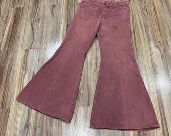 May include: A pair of dusty rose-colored bell-bottom pants featuring a high-waisted design, a button closure at the waistband, and two front pockets. The pants have a wide-leg flare silhouette that widens significantly from the knee down.