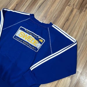 May include: A blue sweatshirt with white stripes on the sleeves and a yellow and white Minnesota license plate graphic that says "SWEDE Land of 10,000 Swedes".