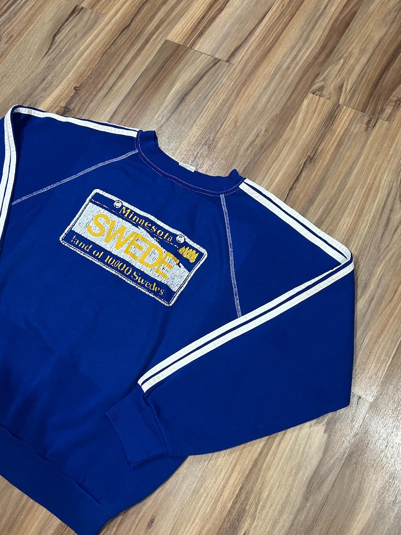 May include: A blue sweatshirt with white stripes on the sleeves and a yellow and white Minnesota license plate graphic that says "SWEDE Land of 10,000 Swedes".