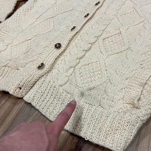 May include: A cream-colored knitted cardigan with a cable knit pattern and button closure. The cardigan has a slightly distressed look with a small hole near the bottom.