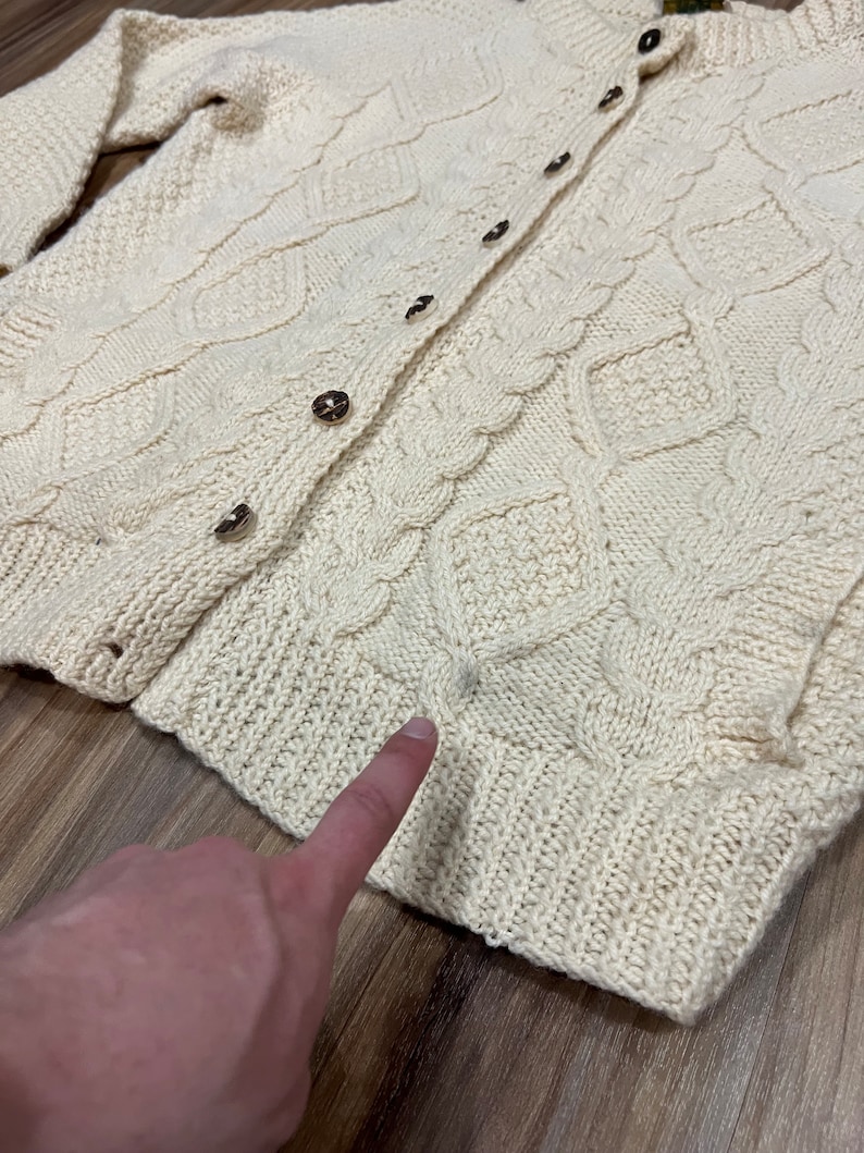 May include: A cream-colored knitted cardigan with a cable knit pattern and button closure. The cardigan has a slightly distressed look with a small hole near the bottom.