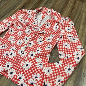 May include: A red and white gingham blazer with a daisy floral pattern. The blazer has a collared neckline, button closure, and long sleeves. The daisies have white petals and navy blue centers.