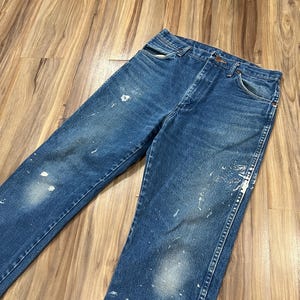 Pu&ograve; includere: Un paio di jeans in denim blu a lavaggio medio con taglio a vita alta, design classico a cinque tasche e chiusura con bottoni. Il tessuto presenta un'estetica vissuta con schizzi di vernice bianca sparsi sulla parte anteriore.