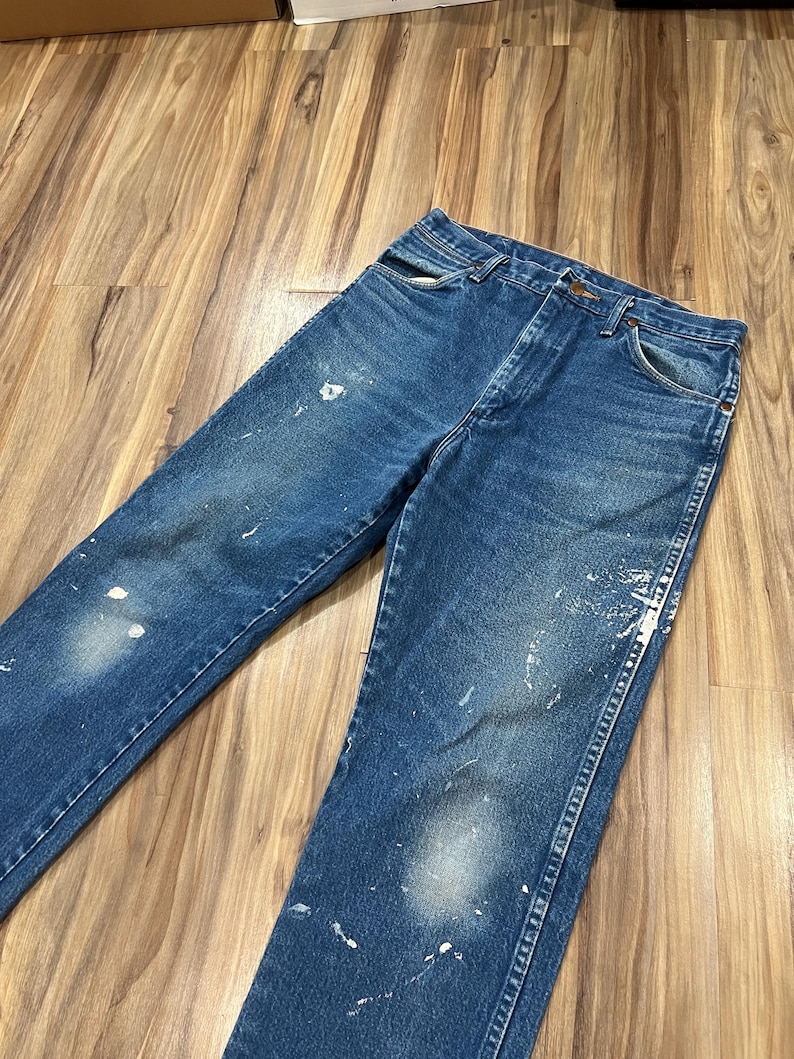 Pu&ograve; includere: Un paio di jeans in denim blu a lavaggio medio con taglio a vita alta, design classico a cinque tasche e chiusura con bottoni. Il tessuto presenta un'estetica vissuta con schizzi di vernice bianca sparsi sulla parte anteriore.