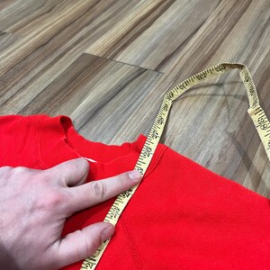 May include: A bright red sweatshirt is measured with a yellow measuring tape. The tape shows a measurement of approximately 23 inches. The sweatshirt is made of a soft material and has a crew neck.