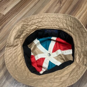 May include: A tan bucket hat with a multi-colored interior lining featuring red, teal, and tan panels separated by white fabric strips. A small blue rectangular tag with the text Pen West is sewn into the inner black band of the hat.