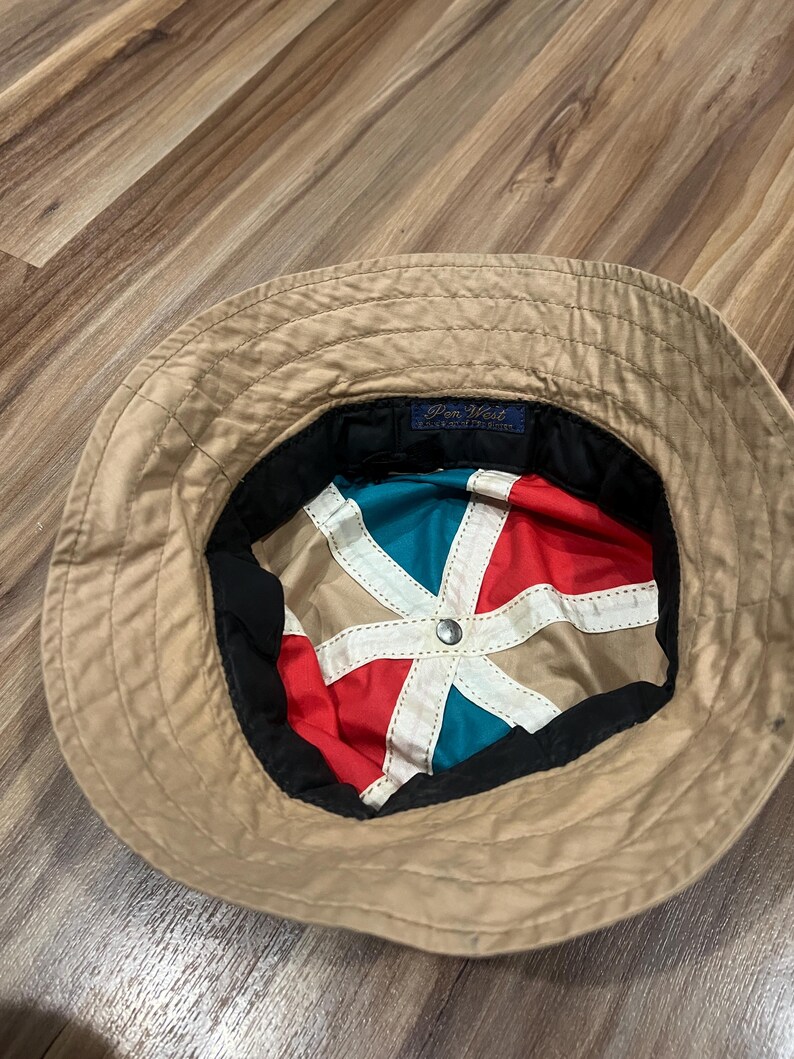 May include: A tan bucket hat with a multi-colored interior lining featuring red, teal, and tan panels separated by white fabric strips. A small blue rectangular tag with the text Pen West is sewn into the inner black band of the hat.