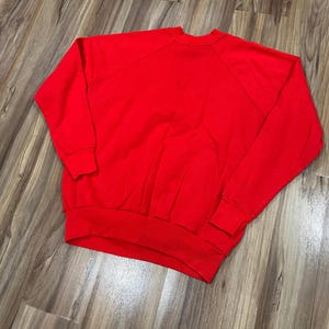 May include: A solid red sweatshirt with a crew neck and long sleeves. The sweatshirt has ribbed cuffs and a ribbed hem. The raglan sleeves extend from the collar to the underarm. The garment is made of a soft material.