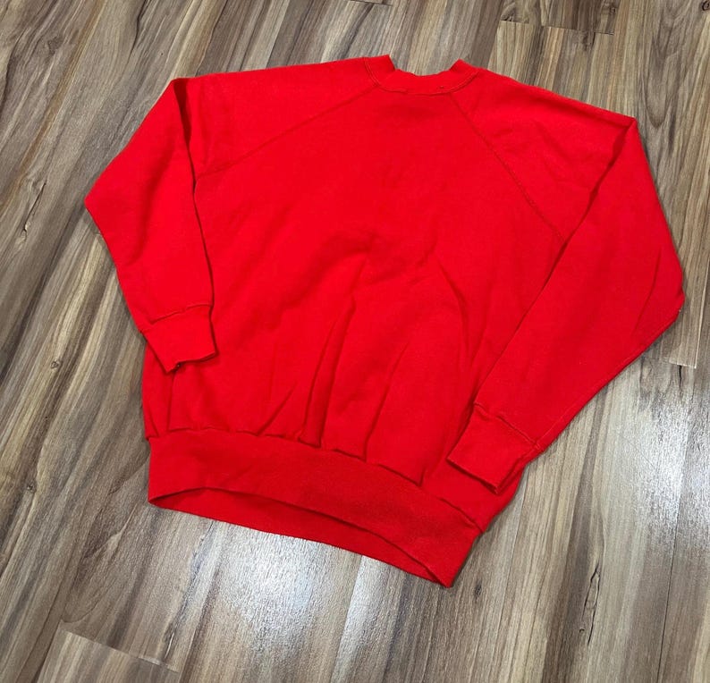 May include: A solid red sweatshirt with a crew neck and long sleeves. The sweatshirt has ribbed cuffs and a ribbed hem. The raglan sleeves extend from the collar to the underarm. The garment is made of a soft material.