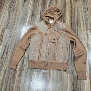May include: A tan zip-up hoodie with a brown suede front panel. The jacket features a hood, long sleeves, and two front pockets. The suede panel has decorative stitching.