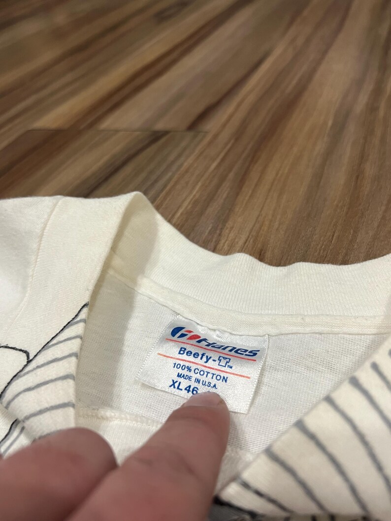 May include: Close-up of a white t-shirt tag. The tag reads "Gildan Beefy-T", "100% Cotton", and "Made in U.S.A.". The size is XL 46.