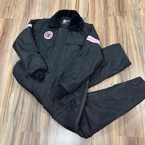 May include: Black snowsuit with a faux fur collar and pink stripes on the sleeves. The snowsuit has a quilted design, a button-up front, and a circular pink emblem on the chest. The pants are attached to the jacket.