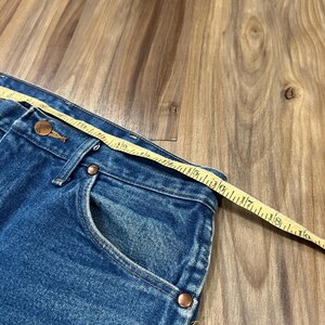 Pu&ograve; includere: Jeans in denim blu con bottone e rivetti color rame, con un metro a nastro giallo posizionato lungo la vita che mostra una misura di circa 46 cm dalla cucitura laterale al bottone centrale anteriore.