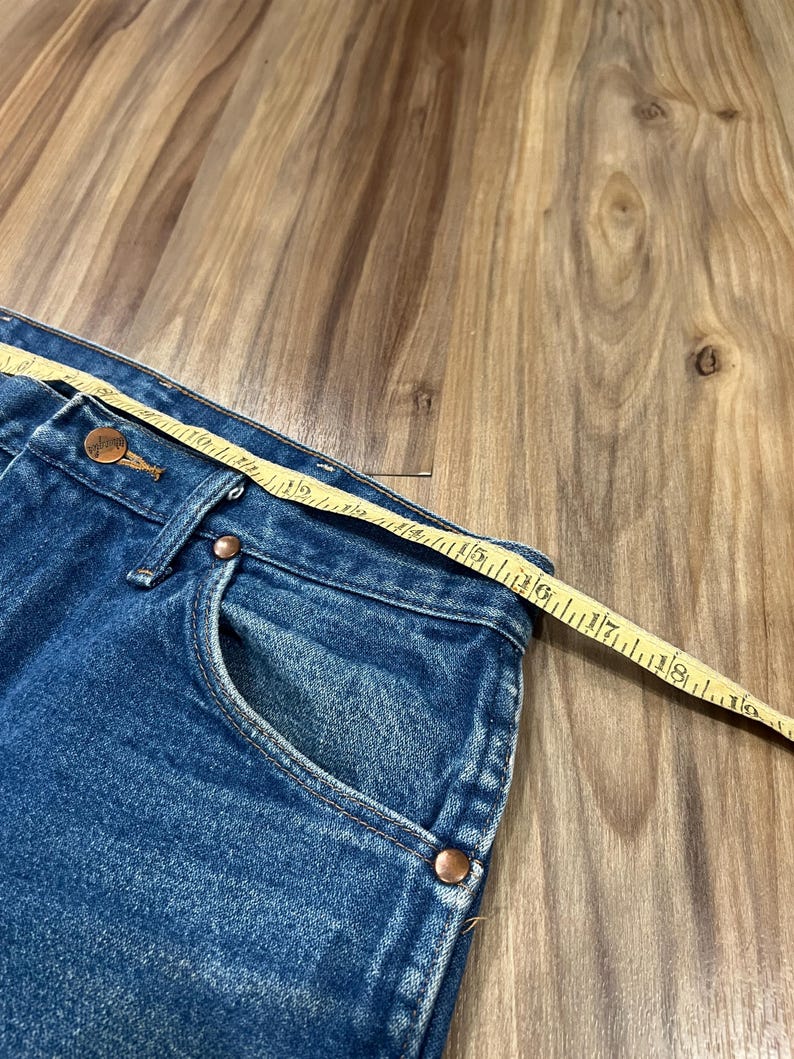 Pu&ograve; includere: Jeans in denim blu con bottone e rivetti color rame, con un metro a nastro giallo posizionato lungo la vita che mostra una misura di circa 46 cm dalla cucitura laterale al bottone centrale anteriore.