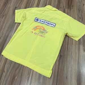 May include: Bright yellow short-sleeved shirt with a collar. The back of the shirt features the "Eastern" logo in a blue rectangle and a graphic of an airplane with the text "Fly Eastern".