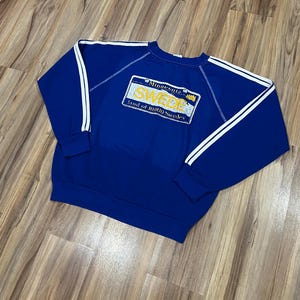 May include: A blue sweatshirt with white stripes on the sleeves. The sweatshirt has a yellow and white graphic of a Minnesota license plate that says "SWEDE Land of 10,000 Swedes".