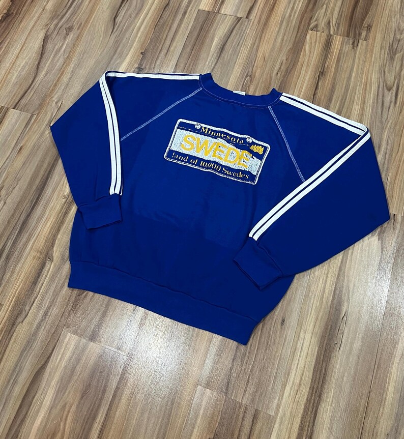 May include: A blue sweatshirt with white stripes on the sleeves. The sweatshirt has a yellow and white graphic of a Minnesota license plate that says "SWEDE Land of 10,000 Swedes".