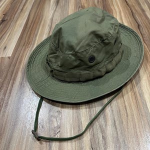 Peut inclure: Un chapeau de brousse vert olive doté d'un large bord, d'une bande en tissu autour de la calotte et d'un œillet d'aération en métal noir de chaque côté. Le chapeau comprend une mentonnière réglable avec un curseur en plastique.