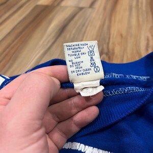 May include: A blue sweatshirt with white and yellow lettering that reads "Minnesota" on the front. The care label is visible and reads "Machine wash separately warm, tumble dry low, no bleach, do not iron or dry clean emblem."