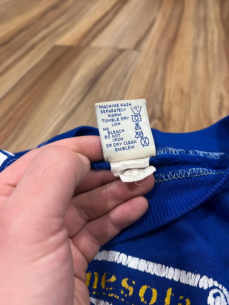 May include: A blue sweatshirt with white and yellow lettering that reads "Minnesota" on the front. The care label is visible and reads "Machine wash separately warm, tumble dry low, no bleach, do not iron or dry clean emblem."