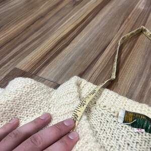 May include: A cream-colored knitted sweater with a brown button and a measuring tape across the top. The sweater has a label that says "42".