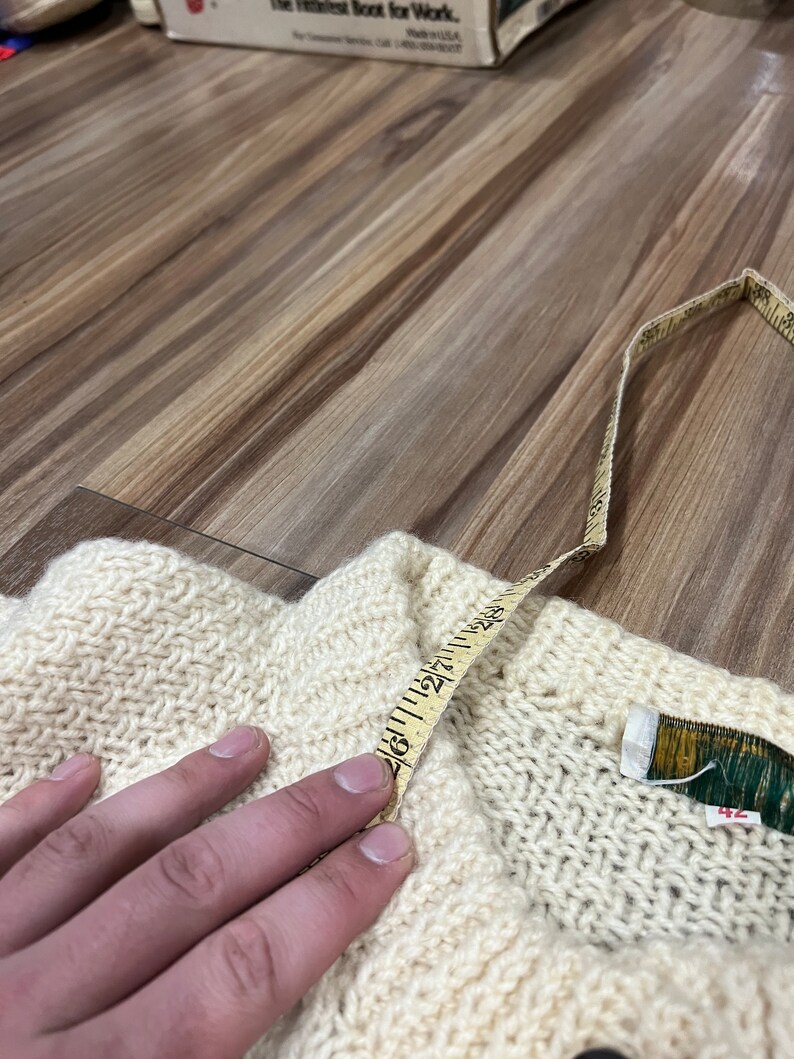 May include: A cream-colored knitted sweater with a brown button and a measuring tape across the top. The sweater has a label that says "42".