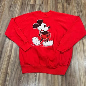 May include: A red sweatshirt featuring a graphic of Mickey Mouse. The cartoon character is depicted in black, white, and red, with a smiling expression. The sweatshirt has long sleeves and a crew neck, suitable for casual wear.