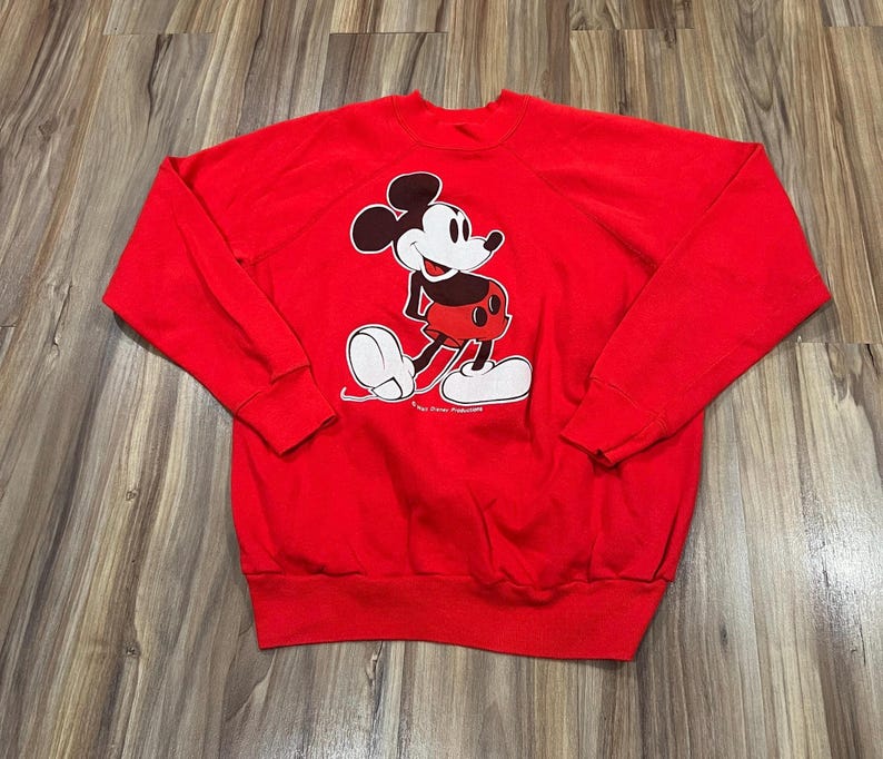 May include: A red sweatshirt featuring a graphic of Mickey Mouse. The cartoon character is depicted in black, white, and red, with a smiling expression. The sweatshirt has long sleeves and a crew neck, suitable for casual wear.