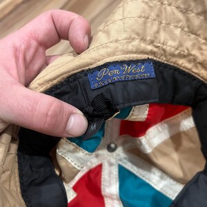 May include: A tan bucket hat with a multi-colored interior featuring red, teal, and white panels. The interior includes a blue woven label that reads Pen West a division of Pendleton and a black size tag with the letter M printed in white.