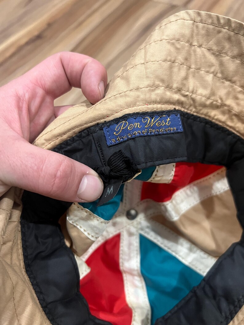 May include: A tan bucket hat with a multi-colored interior featuring red, teal, and white panels. The interior includes a blue woven label that reads Pen West a division of Pendleton and a black size tag with the letter M printed in white.