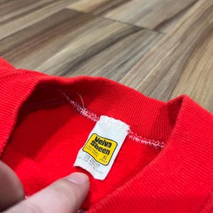 May include: Close-up of a red sweatshirt with a ribbed neckline. A white tag with yellow and black text is visible, reading "Veiva Sheen, Cincinnati, Ohio USA, XL Adult." The tag also lists the fabric content as 50% cotton and 50% acrylic. The sweatshirt is made of a soft, textured material.