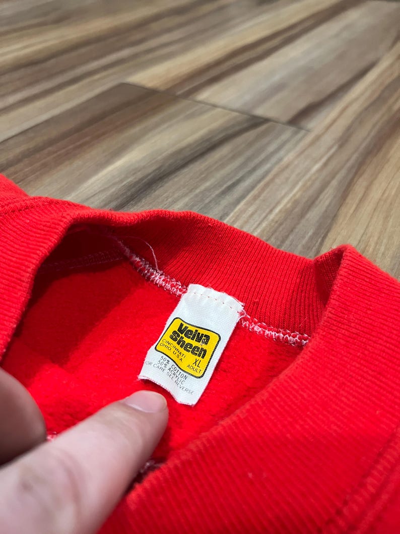 May include: Close-up of a red sweatshirt with a ribbed neckline. A white tag with yellow and black text is visible, reading "Veiva Sheen, Cincinnati, Ohio USA, XL Adult." The tag also lists the fabric content as 50% cotton and 50% acrylic. The sweatshirt is made of a soft, textured material.