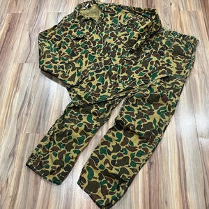 May include: A long-sleeve button-up shirt and matching trousers set featuring a camouflage pattern in shades of olive green, dark brown, and tan. The shirt includes a collar, a front button closure, and two chest pockets with buttoned flaps.