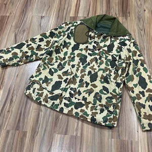 May include: A long-sleeve jacket featuring a camouflage pattern in shades of tan, dark green, and brown. The jacket includes a solid olive green hood, a front button closure, two large chest pockets with flaps, and a small rectangular olive patch.