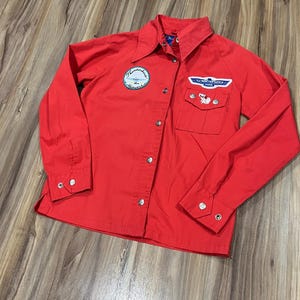 May include: Bright red button-down shirt with long sleeves. Features two patches: one circular with text and a logo, and another rectangular with a logo and a small cartoon cat. The shirt has silver buttons and a collared neckline.