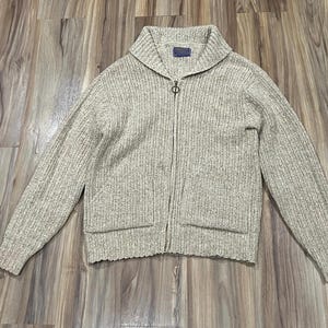 May include: A light beige zip-up cardigan with a ribbed knit texture. The sweater features a shawl collar, two front pockets, and a small label near the collar. The zipper is silver-toned.