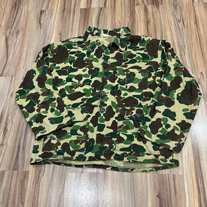 May include: A long-sleeve button-up jacket featuring a camouflage pattern in shades of tan, dark green, and brown. The jacket includes a pointed collar, a single chest pocket on the left side, and two lower front pockets with buttoned flaps.